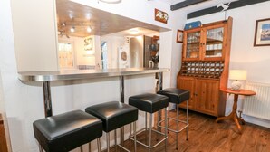 A kitchen with wooden bar stools, a wooden cabinet with glass doors, and a small table with a lamp. - The Cottage (Pwllheli)