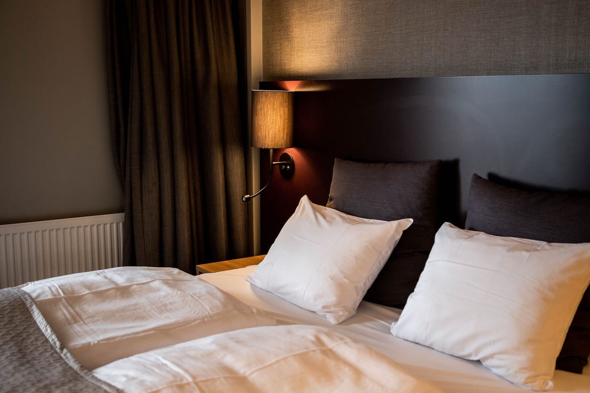 A hotel room with a neatly made bed, a headboard with a light fixture, and a window with curtains.
