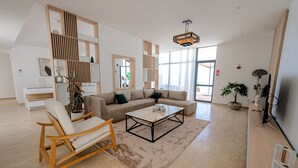 Un salón amplio con un sofá modular grande, una mesa de centro de madera y una silla de madera. Hay un ventanal que ofrece luz natural y vistas al exterior.