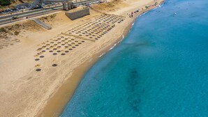 Vista aérea de una playa con hileras de sombrillas y sillas, un mar azul y una carretera con vehículos.