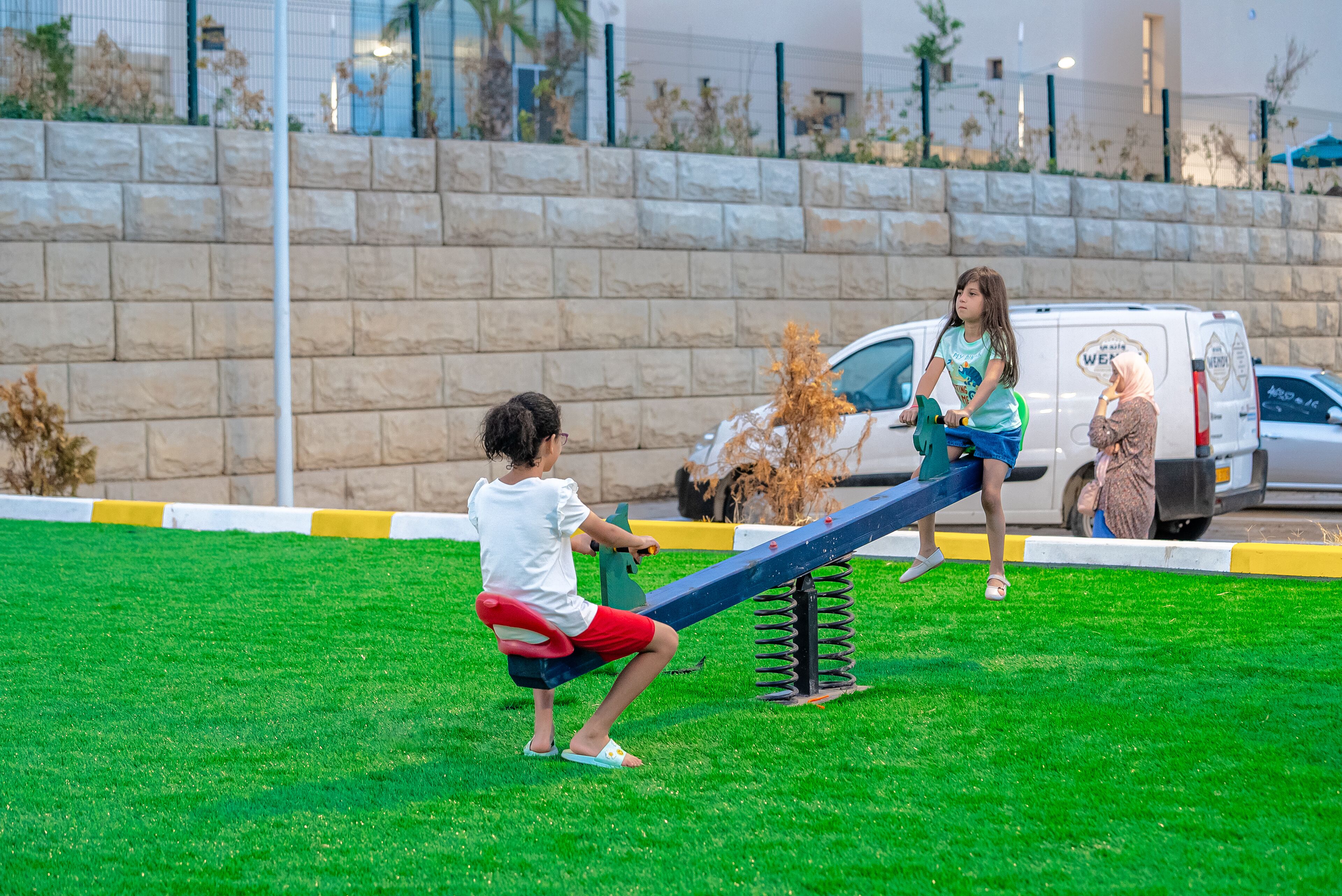 Two children playing on a seesaw in a park with a white van in the background.