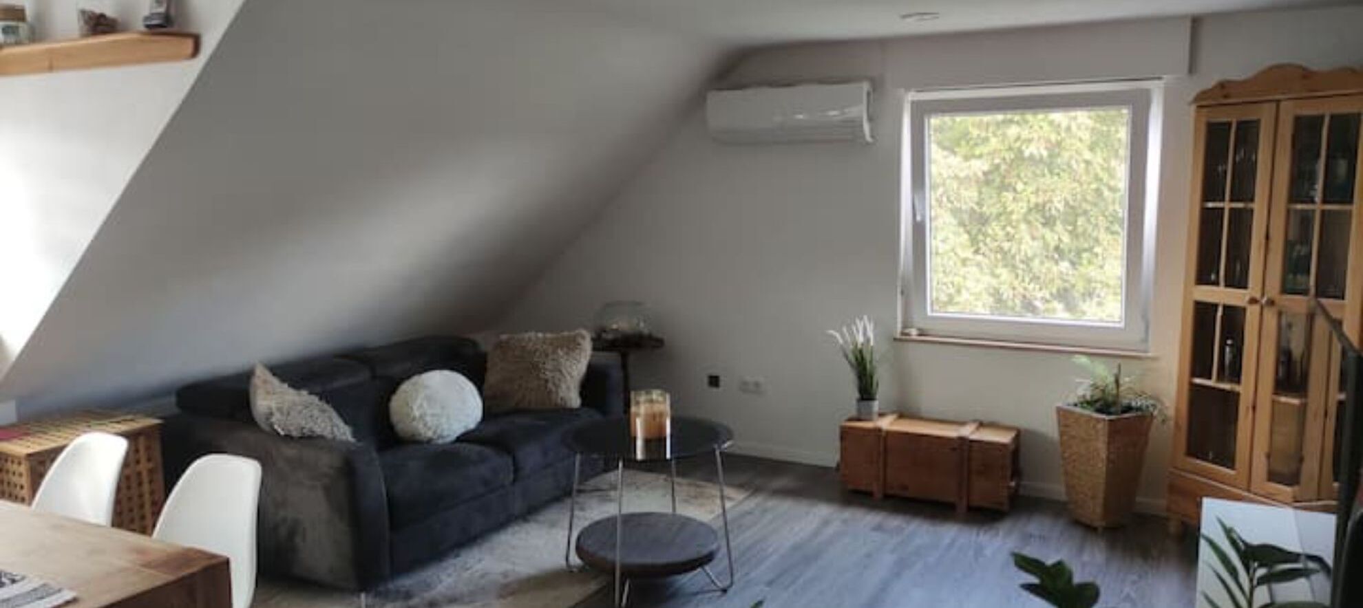 Ein Loft-Zimmer mit einem dunkelgrauen Sofa, einem kleinen runden Tisch und einem Fenster mit Blick auf Bäume.