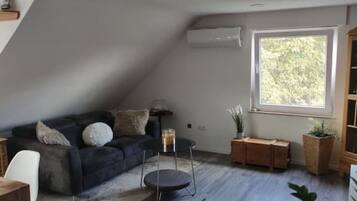 Ein Loft-Zimmer mit einem dunkelgrauen Sofa, einem kleinen runden Tisch und einem Fenster mit Blick auf Bäume.