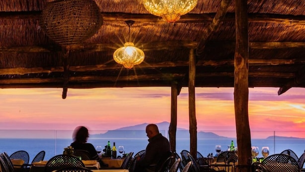 An outdoor dining area with tables and chairs, a thatched roof, and hanging lights.