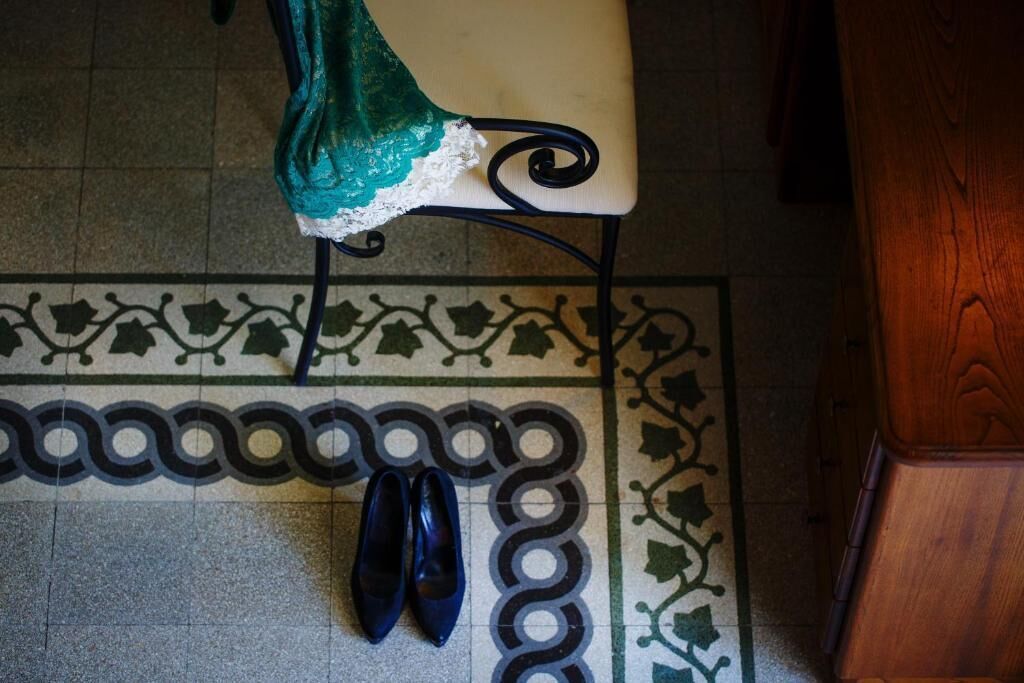 Tuoli, jossa on vihreä kangasverhoilu ja valkoinen tyyny, pari sinisiä kenkiä kuvioiduilla lattialla ja puinen huonekalu.