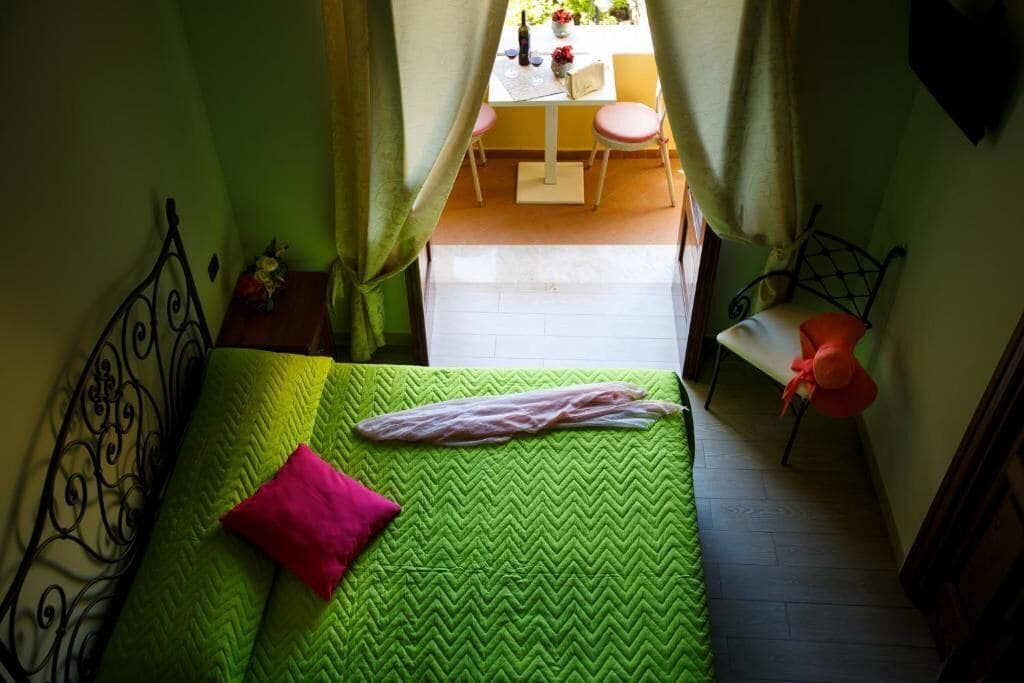 Une petite chambre à coucher, chaleureuse et accueillante, avec une couvre-lit verte, un oreiller rose et une tête de lit en métal. Une petite table avec une chaise est visible à travers une porte, ce qui laisse supposer qu’il s’agit d’un espace partagé.