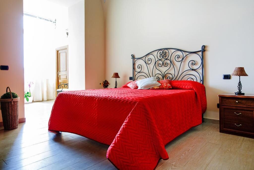 Un dormitorio con estructura de cama metálica, colcha roja y mesita de noche de madera con lámpara.