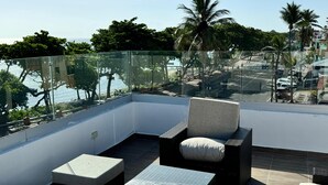 A balcony with a glass railing, a wicker chair, a small table, and a view of a beach and palm trees. - Central Apartment Facing the sea (Puerto Plata)