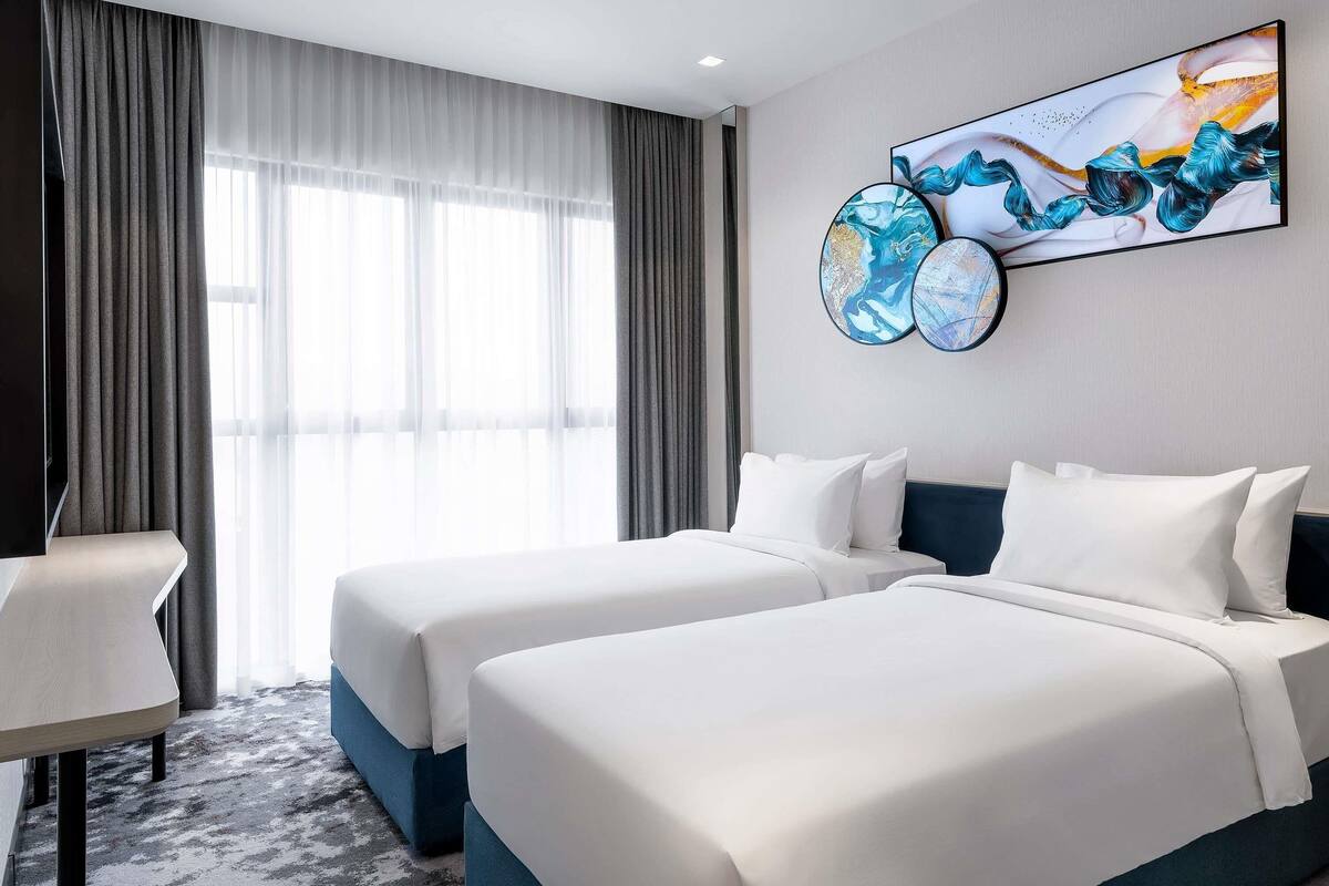 A hotel room with two beds, a desk, and a large window with sheer curtains.