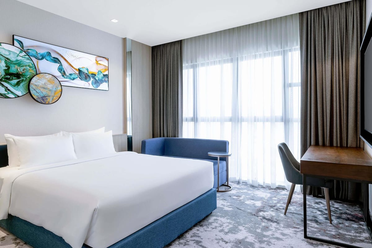 A hotel room with a large bed, a desk, and a chair. There is a large window with curtains, and artwork on the wall.