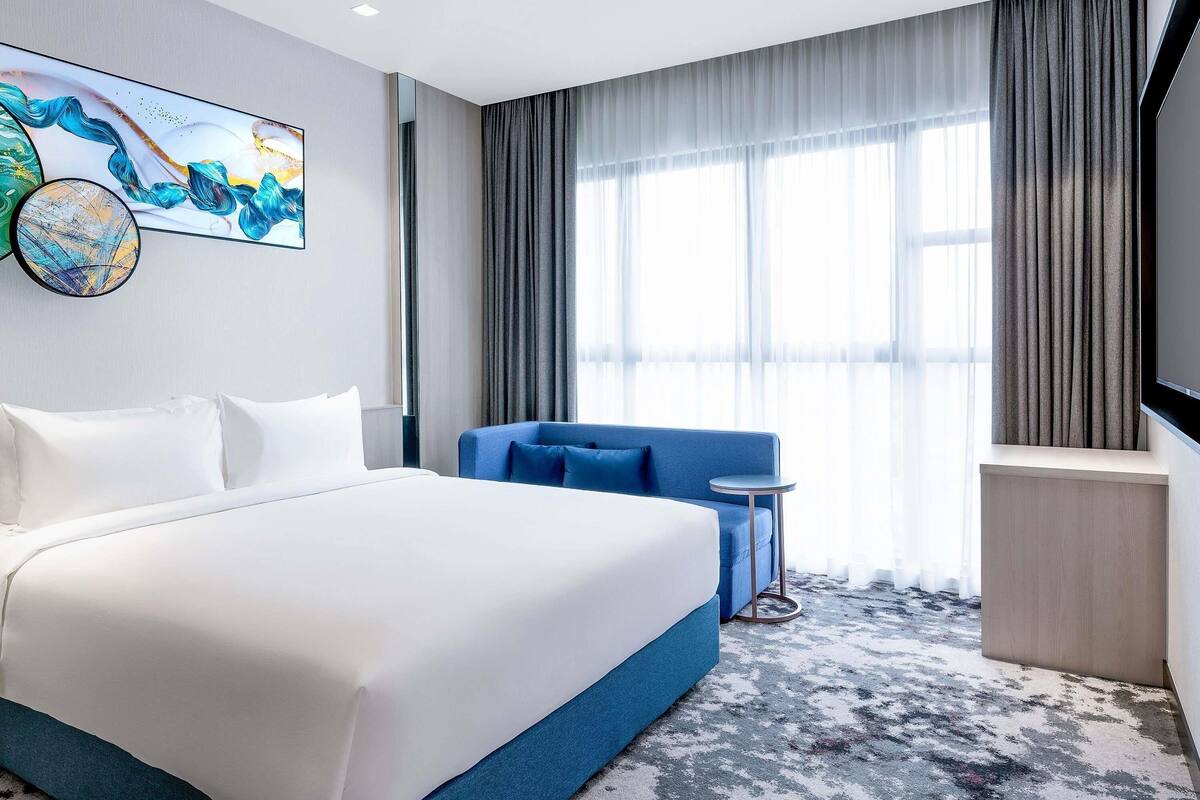 A hotel room with a large bed, a blue sofa, a small table, and a large window with curtains.
