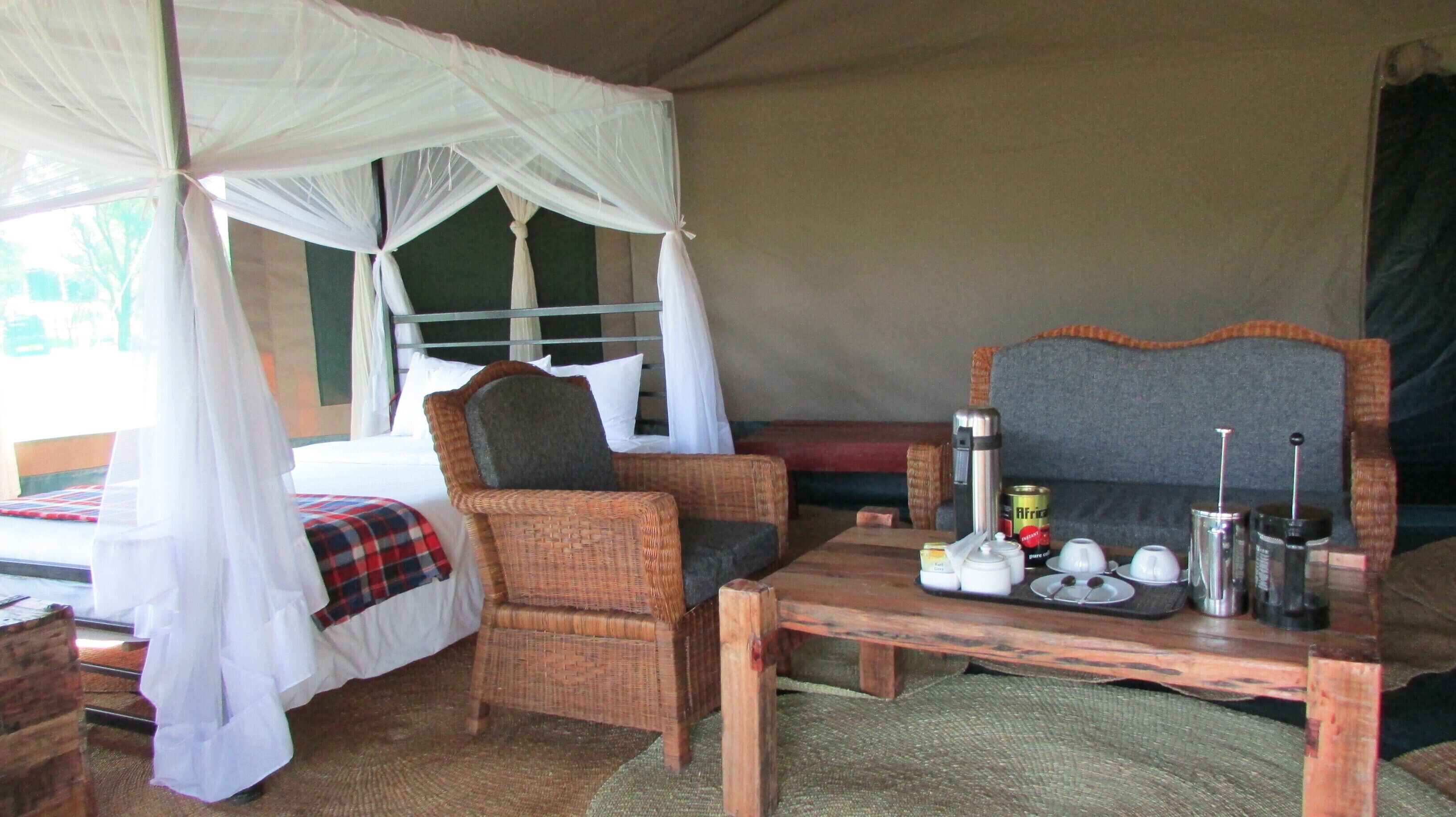 Una habitación con carpa, una cama, una silla de mimbre, una mesa con tazas y un termo, y una ventana con cortinas.