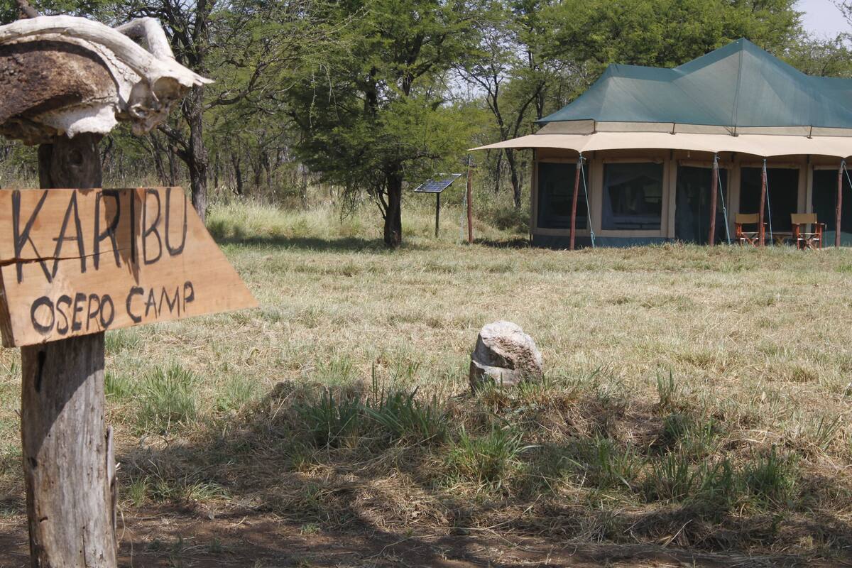 A sign for Karibu Osero Camp in a natural setting with a tent structure in the background.