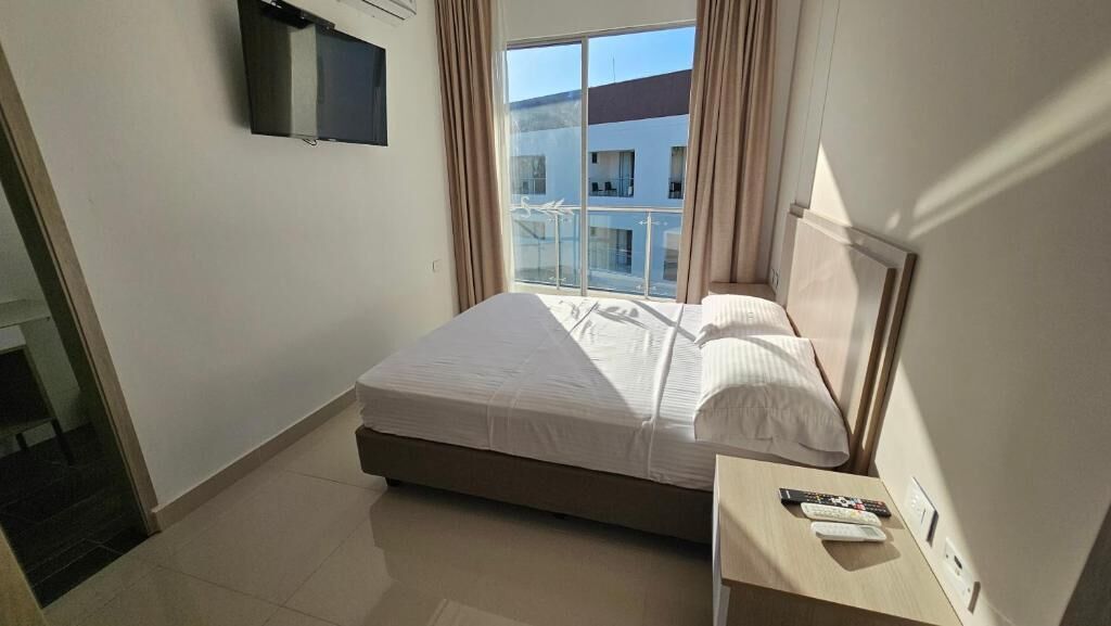 A hotel room with a bed, a TV mounted on the wall, a bedside table with a remote control, and a view of the outside balcony.