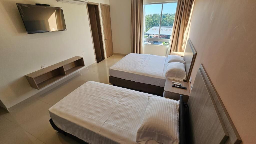 A hotel room with two beds, a TV mounted on the wall, a wooden shelf, and a window with curtains.