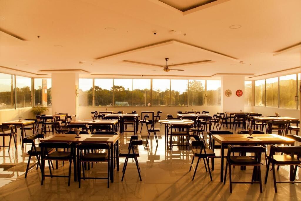 A spacious dining area with multiple tables and chairs, large windows offering a view of the outdoors, and a ceiling fan.