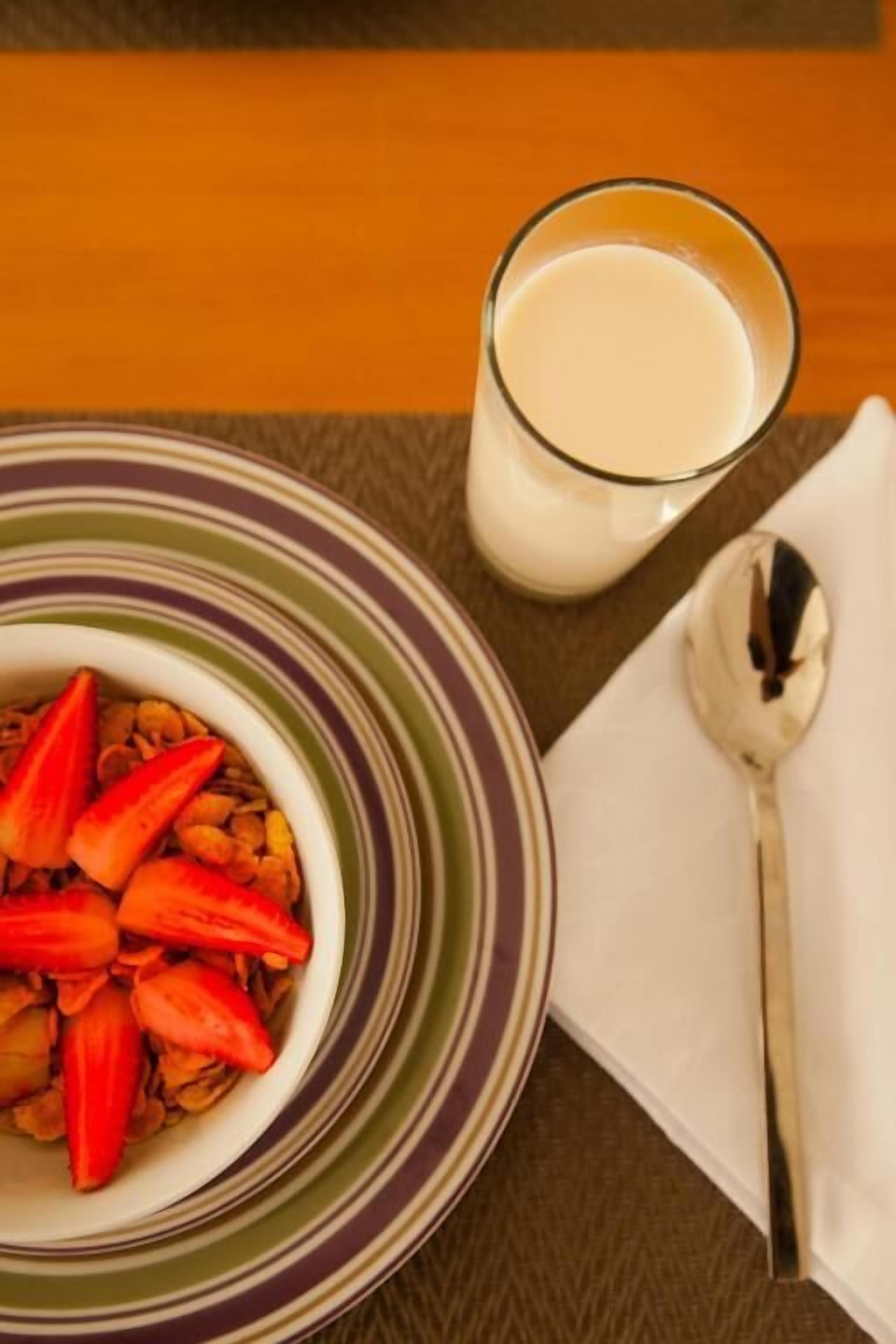 Een ontbijtsetting met een kom ontbijtgranen, belegd met aardbeien, een glas melk en een lepel op een servet.