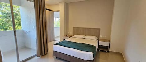 A modern hotel room with a large bed, a nightstand, a sliding glass door to a balcony, and a view of greenery outside.