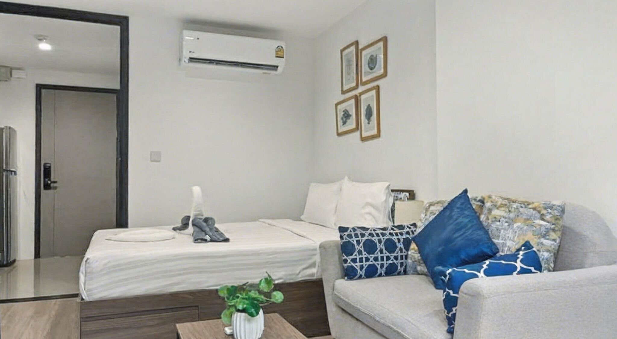 A hotel room with a bed, a sofa, a small table with a plant, and an air conditioning unit on the wall.