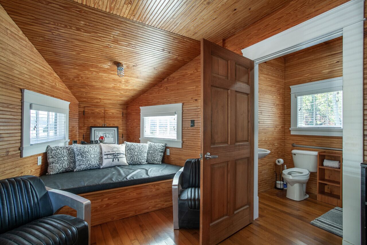 Un intérieur de chalet chaleureux avec un plafond en bois, un banc intégré avec des coussins, une porte donnant sur une salle de bain et une fenêtre avec des stores.