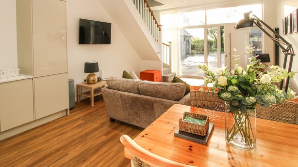 A modern living room with a sofa, a wooden dining table, a TV mounted on the wall, and a staircase leading to an outdoor area. - The Lawley (Shrewsbury)
