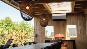 Een terras met houten tafels en stoelen, een barbecue en hangende lampen.