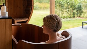 Une personne dans une baignoire en bois, avec vue sur un jardin à l’extérieur.