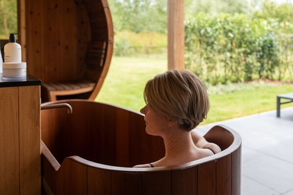 Une personne dans une baignoire en bois, avec vue sur un jardin à l’extérieur.