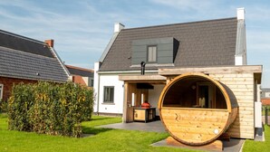 A modern house with a large wooden barrel sauna in the front yard.