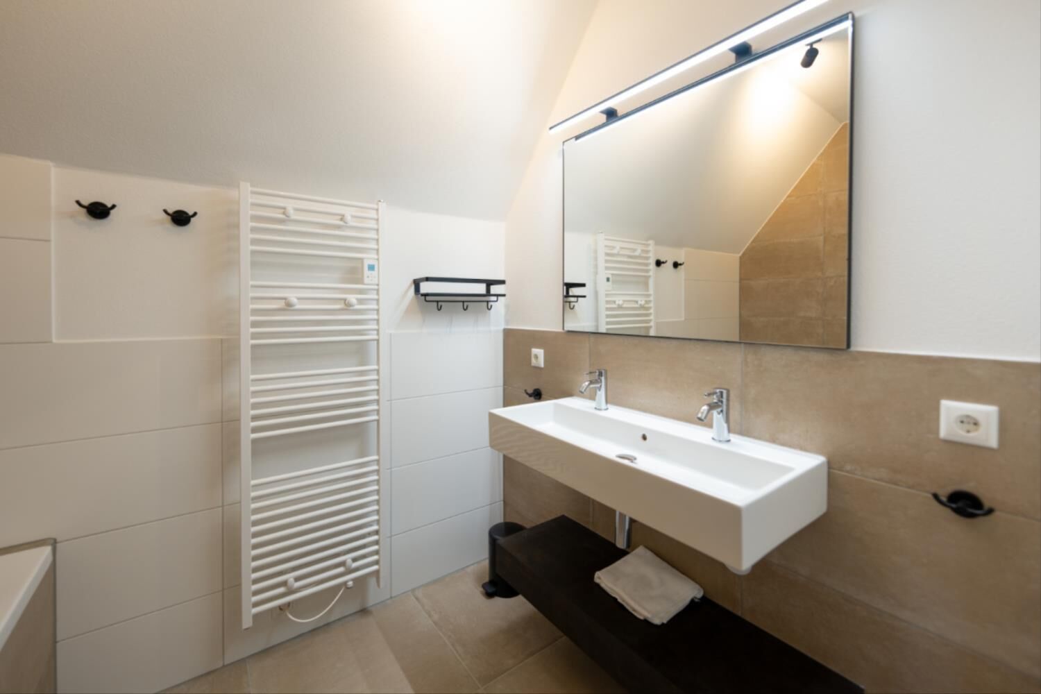Une salle de bain moderne avec un lavabo blanc, deux robinets, un miroir doté d’un éclairage intégré, un porte-serviettes et une serviette posée sur le sol.