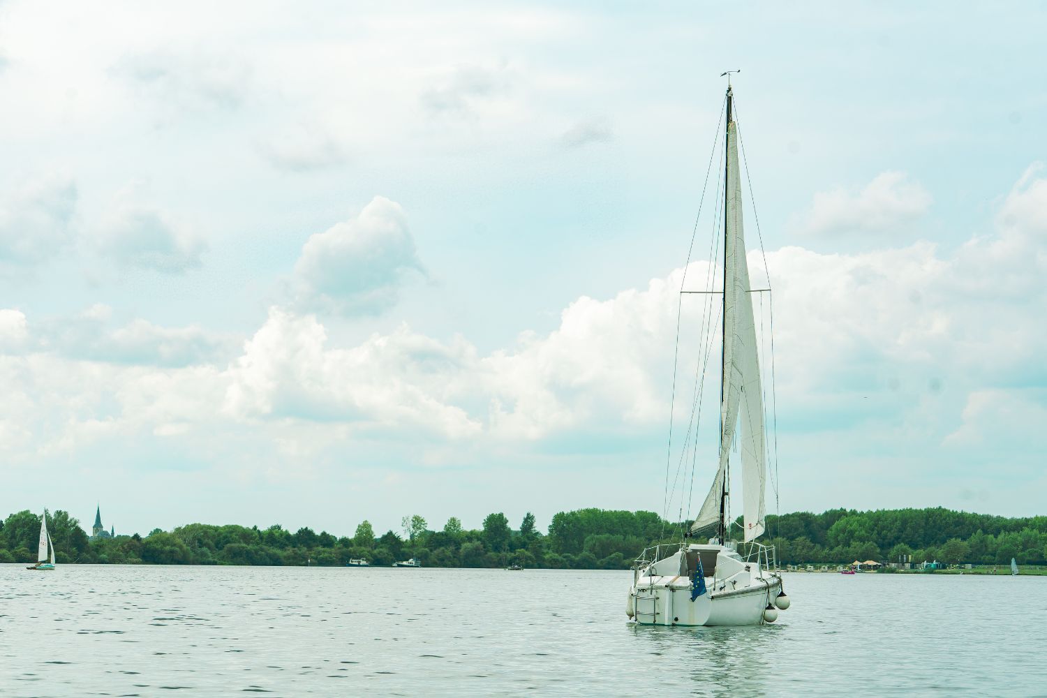 Een zeilboot op kalm water, met een heldere lucht en een bebost oever in de verte.