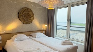 Ein Hotelzimmer mit einem großen Bett, einer Wanddekoration, einer Nachttischlampe und Blick auf den Strand durch das Fenster.