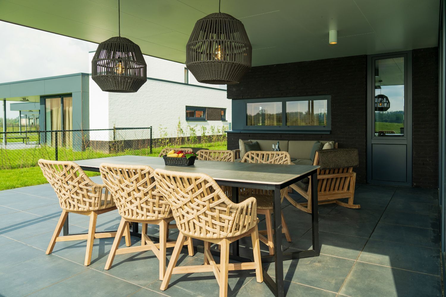 An outdoor dining area with wicker chairs and a table, surrounded by a modern building with large windows and a view of the countryside.