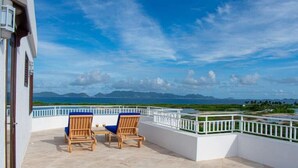 Une terrasse avec deux chaises en bois et une table, d’où l’on peut admirer un paysage côtier avec des montagnes et une étendue d’eau.