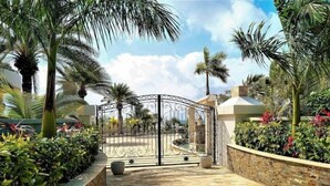 A gated entrance with a stone pathway leading to a building, surrounded by palm trees and various plants.