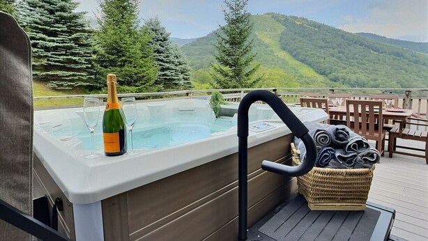 A hot tub with a bottle of champagne and glasses, a wicker basket with towels, and a view of mountains. - SlopeSide- Pool, Grill, Hot tub, Views! (Killington)