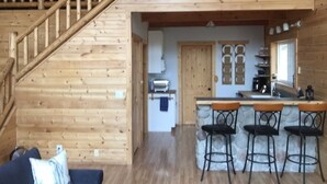 Interior de una cabaña de madera con una escalera que lleva a un dormitorio en el altillo, una cocina con barra y una sala de estar con sofá.