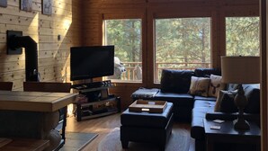 Interior acogedor de una cabaña con techo de madera, ventanas amplias, televisor, sofá y chimenea.