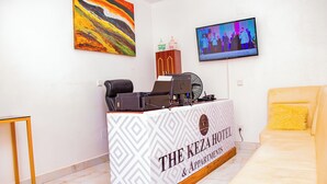 A reception desk with a logo, a television mounted on the wall, and a sofa with a yellow pillow. - The Keza Hotel and Apartments (Kigali)