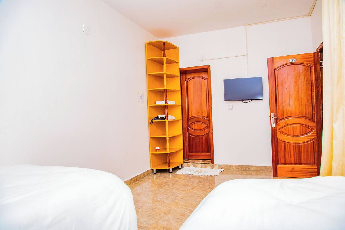 A hotel room with two beds, a wooden wardrobe, a television mounted on the wall, and a yellow shelving unit.