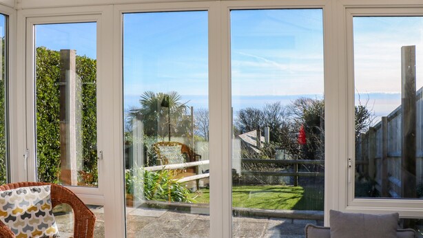 A conservatory with large glass doors, wicker furniture, and a view of a garden and distant landscape. - Saltwater Cottage (Ventnor)