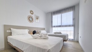 Una habitación de hotel moderna con una cama grande, otra más pequeña, una mesita de noche y un ventanal amplio.