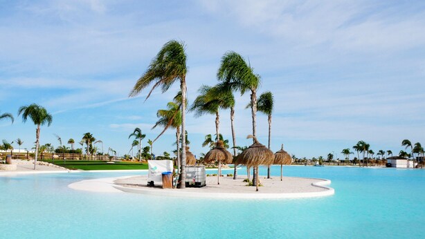Área de piscina en un resort, con palmeras, una pequeña isla y una silla de descanso, bajo un cielo azul y despejado.