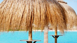 Una sombrilla de techo de paja, dos mesas de madera y una playa arenosa con agua azul cristalina al fondo.