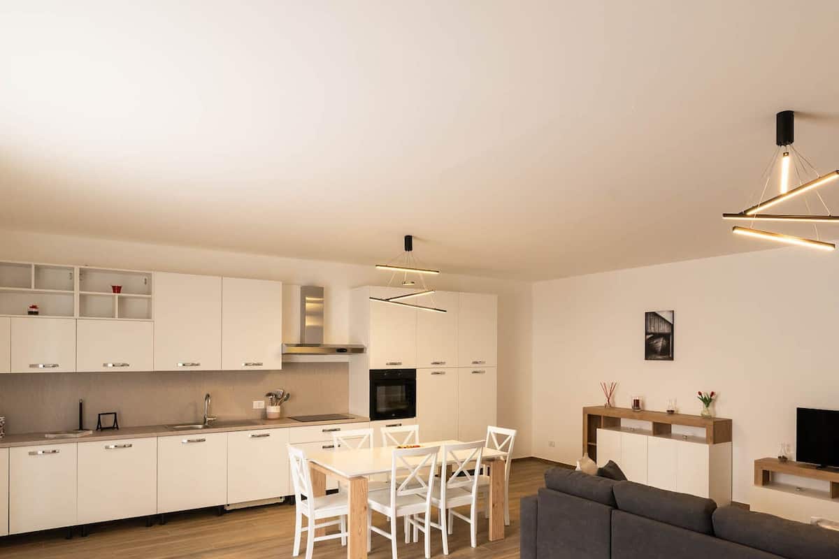A modern kitchen with white cabinets, a dining area with a white table and chairs, and a living room with a gray sofa and a TV.
