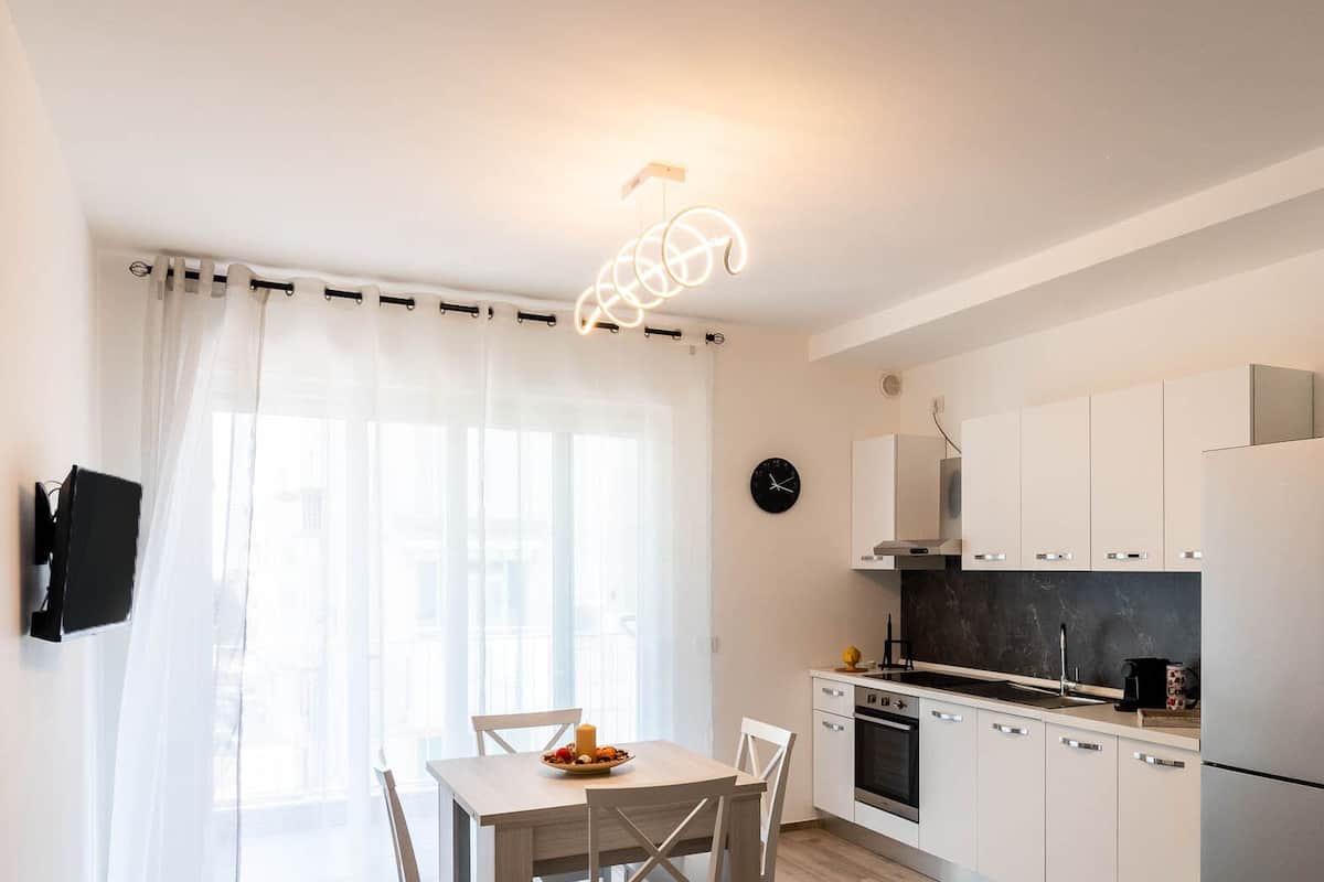 A modern kitchen with white cabinets, a black stove, and a refrigerator. There is a dining table with chairs and a candle on it. A clock is on the wall.