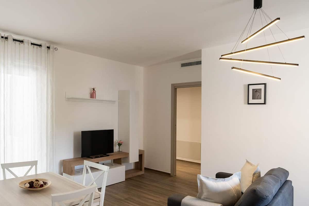 A modern living room with a TV, a dining area with a table and chairs, and a sofa with cushions.