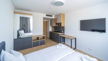 A modern hotel room with a bed, a desk, a kitchenette, and a TV.