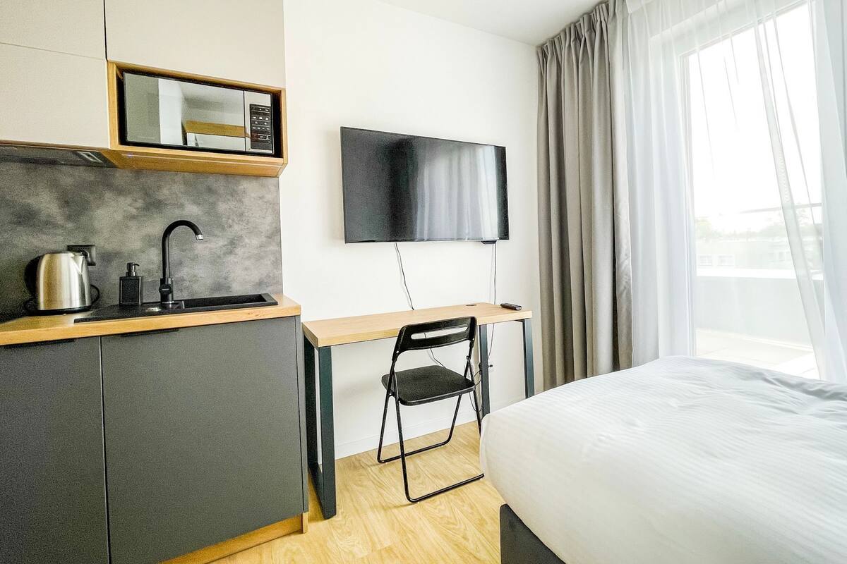 Et moderne hotelværelse med tekøkken, fladskærms-TV, seng og et stort vindue med gardiner.