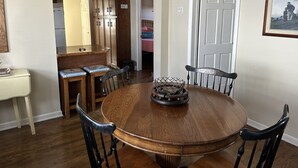 Un comedor con mesa y sillas de madera, una cocina al fondo y una puerta con un cuadro enmarcado.